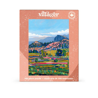 The Villager Puzzles “Wine Country | 500 Piece Jigsaw Puzzle” features a vibrant painted landscape of fields, hills, trees, and distant mountains beneath a blue sky, with artwork inspired by Amy Dixon.