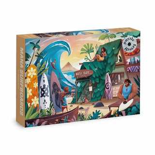 A Mana Surfboards | 500 Piece Jigsaw Puzzle box featuring an illustrated tropical surf shop scene with surfboards, a large breaking wave, and several characters.
