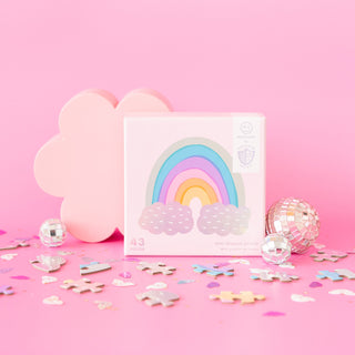 With a pink background, pastel rainbow "Over the Rainbow | 43 Piece Jigsaw Puzzle" by Jollity & Co., pink cloud decor, disco balls, confetti, and scattered mini pieces—it's a whimsical party favor.