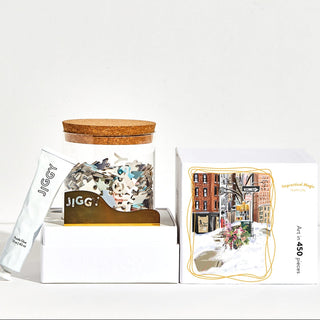 A reusable glass jar with a cork lid holds puzzle pieces, a tube of Jiggy Puzzles glue, a gold tool, and packaging featuring the Impractical Magic | 450 Piece Jigsaw Puzzle by Jiggy Puzzles.