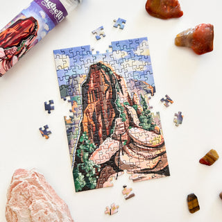 Spaghetti Tree’s Angels Landing 150 Piece Jigsaw Puzzle shows a rocky mountain scene in progress, surrounded by loose pieces, a puzzle tube, and stones on a white surface.