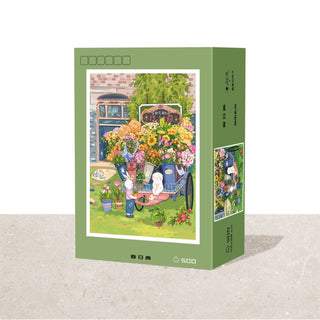 The Ber Spring Sun Green 500 Piece Jigsaw Puzzle comes in a green box featuring a flower cart with vibrant flowers, garden tools, and a cat. Finished puzzle size: 15.7 x 16.5 inches.