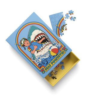 A partially assembled Stay Positive | 99 Piece Jigsaw Puzzle by Ohh Deer, featuring a box with a cartoon diver, large shark, beach ball, and the words “STAY POSITIVE” on a blue background. Nearby, loose pieces from this dark humor puzzle inspired by Steven Rhodes are scattered.