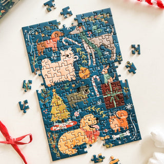 The partially completed Merry Woofmas | 150 Piece Jigsaw Puzzle by Spaghetti Tree features adorable animal illustrations, a Christmas tree, and festive decorations on a white surface. Loose pieces from this charming portable puzzle are scattered around, making it perfect for cozy holiday moments anywhere.