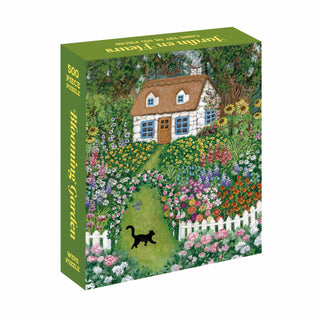 The WEVE Blooming Garden 500 Piece Jigsaw Puzzle features a velvet touch box with an illustration of a cottage, a vibrant garden, and a black cat strolling by a white picket fence.