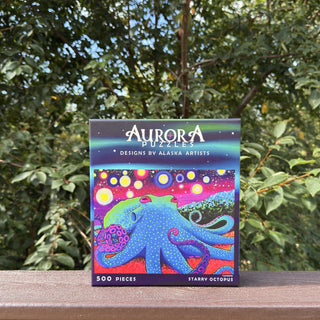 The Aurora Puzzles Starry Octopus | 500 Piece Jigsaw Puzzle features vibrant giant Pacific octopus artwork, making it a perfect choice for puzzle lovers. Box shown on a wooden railing with trees in the background.