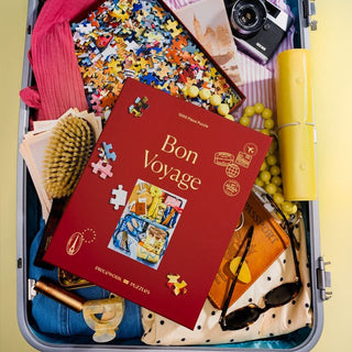 An open suitcase with a Piecework Puzzles Bon Voyage | 1,000 Piece Jigsaw Puzzle box, assorted puzzle pieces, clothes, a hairbrush, scarf, jewelry, camera, sunglasses, book, and travel accessories.