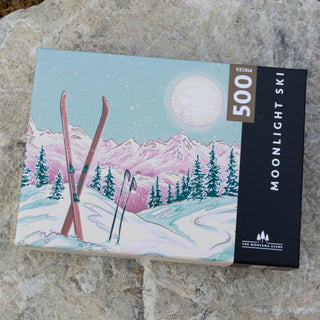 The Montana Scene's Moonlight Ski | 500 Piece Jigsaw Puzzle shows skis and poles in the snow with mountains and a large moon, capturing a peaceful Montana setting. The box is pictured on a rocky surface.
