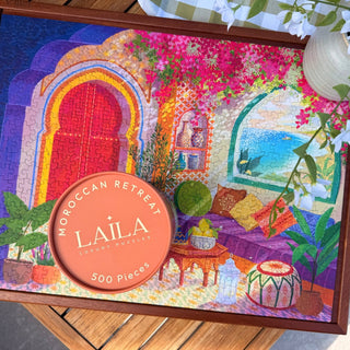 The Moroccan Retreat | 500 Piece Jigsaw Puzzle by Laila Luxury Puzzles features a vibrant Moroccan-inspired courtyard, lush plants, colorful cushions, and elegant arches for an immersive puzzle experience.