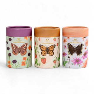 The Flutter Friends set by 1canoe2 includes three 100-piece jigsaw puzzles, each featuring cylindrical containers adorned with floral and butterfly artwork—ideal for nature enthusiasts—and topped with uniquely colored lids in purple, brown, and peach against a white background.