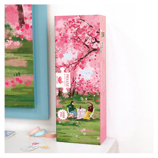 A Momibook "Under the Cherry Tree" 1,000 piece jigsaw puzzle box stands upright on a shelf next to a framed picture, featuring two people having tea beneath cherry blossom trees in a park.