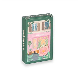 A rectangular green Trevell box with an illustrated storefront labeled "Manuka" and pink flowers contains the charming Manuka | 99 Piece Jigsaw Puzzle.