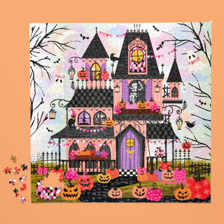The WerkShoppe Boohemian Mansion 1,000 Piece Jigsaw Puzzle features a whimsical haunted house adorned with pumpkins, bats, and flowers on an orange background, with a few loose pieces scattered beside the completed puzzle.