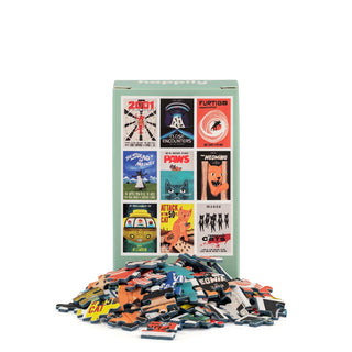 A box of Happily Jigsaw Puzzles’ “Movie Cats | 99 Piece Mini Jigsaw Puzzle”—featuring illustrated sci-fi and horror movie posters with a feline twist—stands behind a pile of vibrant pieces, perfect for cat lovers seeking a quirky puzzle experience.