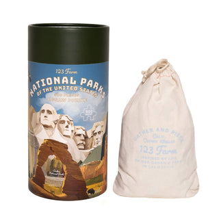 A cylindrical box labeled "National Parks | 500 Piece Jigsaw Puzzle" by 123 Farm, featuring sites like Grand Canyon National Park, stands beside a drawstring cloth bag with blue text.