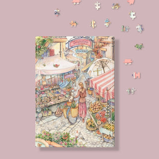 The Puzzle Theory’s Market Day | 896 Piece Jigsaw Puzzle by Kim Jacobs features a bustling outdoor market scene with stalls, produce, and a woman with a bicycle; loose eco-friendly pieces are shown on a pink background.