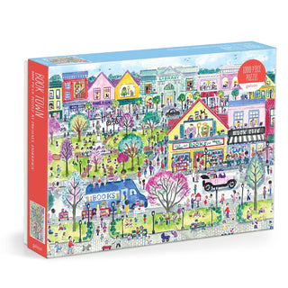 The Michael Storrings Book Town 1,000 Piece Jigsaw Puzzle by Galison features a vibrant illustrated town with bookstores, a library, and people enjoying outdoor activities in a lively park setting.