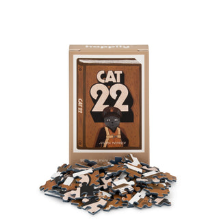A box labeled "Cat 22 | 99 Piece Mini Jigsaw Puzzle" by Happily Jigsaw Puzzles features Nia Gould’s illustrated cover and a character, shown with scattered mini jigsaw pieces.