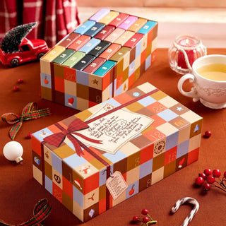 Two rectangular, multi-colored Trevell Advent Calendar Puzzle No. 5 boxes from Trevell sit on a brown table, surrounded by a cup of tea, berries, ribbon, Christmas ornaments, and festive decor—each box containing 24 mini 99-piece jigsaw puzzles to make your Christmas countdown extra special.