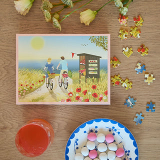 A ViSSEVASSE "Summer" 300 piece jigsaw puzzle lies on a table with a postcard of two cyclists by the sea, surrounded by flowers, near a red drink and a plate of pink and white sweets.