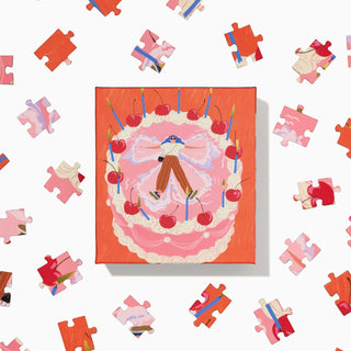 The Cake Angel 100 Piece Jigsaw Puzzle by Ordinary Habit features a cake, candles, cherries, and a central figure on a clean white background. Made from recycled materials, this square mini puzzle is perfect for a charming challenge.