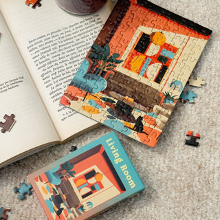 A partially completed Minizz Living Room | 99 Piece Jigsaw Puzzle rests on the carpet near its box and an open book, with scattered pieces evoking the charm of a Parisian interior design.