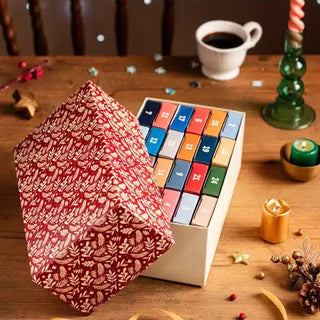 The Trevell Advent Calendar No. 2 by Trevell, featuring 24 mini 99-piece jigsaw puzzles in numbered compartments, is partially opened on a wooden table among coffee, a candle, and decor for a cozy winter scene.