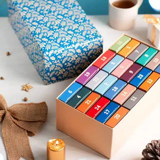 The Trevell Advent Calendar No. 1 by Trevell, featuring 24 colorful drawers each with a 99-piece jigsaw puzzle, sits next to a blue patterned box, ribbon, and festive decorations for a cheerful winter display.