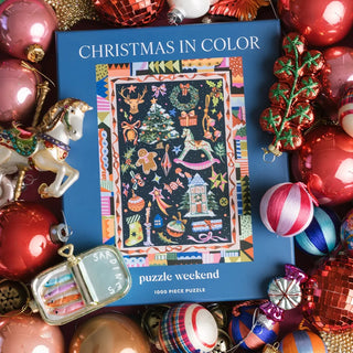 A 1,000-piece Christmas in Color jigsaw puzzle box centered among an assortment of holiday ornaments.