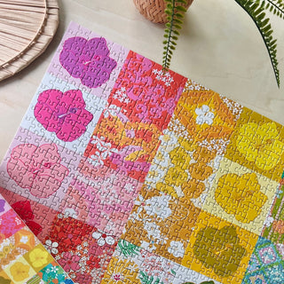 The Anuenue Quilt 1,000 Piece Jigsaw Puzzle by Surf Shack Puzzles, featuring a floral pattern in pink, red, orange, and yellow, is partially completed on a light surface beside a plant and woven decor.