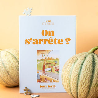 The Jour Férié “On S'arrête? | 500 Piece Jigsaw Puzzle” stands upright with two melons and two puzzle pieces beside it, all set against a peach background. French-made, ribbon cut.