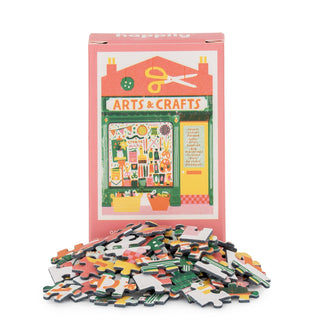 A box labeled "Arts & Craft | 99 Piece Mini Jigsaw Puzzle" by Happily Jigsaw Puzzles, featuring an arts and craft shop illustration, sits behind a pile of assorted mini puzzle pieces made from recycled board.