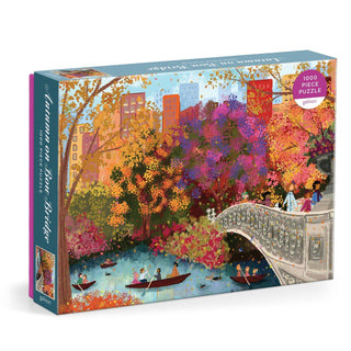 The Galison Joy Laforme Autumn On Bow Bridge 1,000 Piece Jigsaw Puzzle features a vibrant illustration of Central Park’s Bow Bridge in autumn, with colorful trees, people walking, and rowboats on the water.