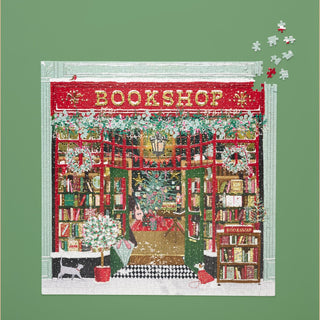 The WerkShoppe Festive Bookshop 1,000 Piece Jigsaw Puzzle features a holiday-themed bookshop with bookshelves and a guitar; a few pieces remain unfinished in the top right corner.