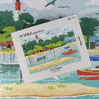 A finished Cap Ferret | 1,000 Piece Jigsaw Puzzle by Aparté with its box, which features the same waterfront scene, displayed atop the completed puzzle.