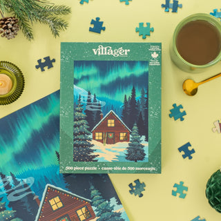 The Villager Puzzles Northern Cabin 500 Piece Jigsaw Puzzle, featuring a snowy forest cabin under the northern lights, sits on a table with scattered pieces, coffee, and Canadian Christmas decor.