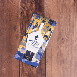 A rectangular Trevell Puzzle in A Bottle Box Set, showcasing a cityscape design, sits on wood. Inside are 12 mini jigsaw puzzles, each with 99 pieces—perfect for puzzle enthusiasts.