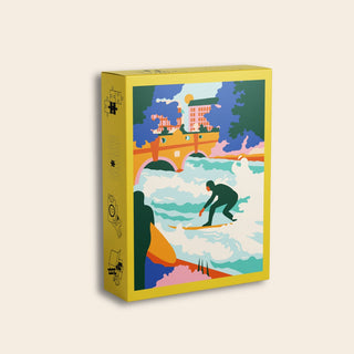 The Eisbach Wave | 99 Piece Jigsaw Puzzle by Das Puzzle Kollektiv comes in a yellow box and features artwork by Katharina Bourjau depicting a person surfing the Eisbach Wave on a river, with a bridge, trees, and buildings in the background. This puzzle is made from recycled paper.