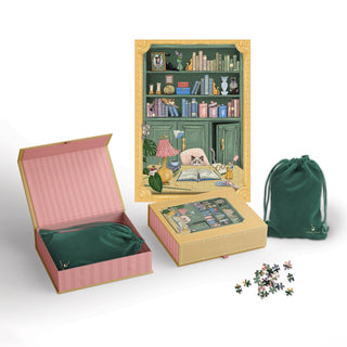 A boxed Cat Library | 500 Piece Jigsaw Puzzle by Good Juju Ink is shown, featuring a completed image of a cat on a bookshelf accented with gold foil details. The set includes a magnetic keepsake box, velvet pouch, and displays the loose puzzle pieces.