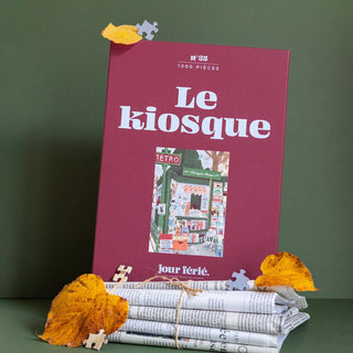 A rectangular Jour Férié Le Kiosque 1,000 Piece Jigsaw Puzzle box with a red top and green bottom depicting a newspaper kiosk, displayed with a rolled newspaper and scattered autumn leaves.