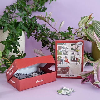 The Fenêtre Sur Cour 100-piece mini puzzle by Pièce Rapportée, made in France, sits open on a vibrant purple background with green potted plants beside its completed image of a decorated room.