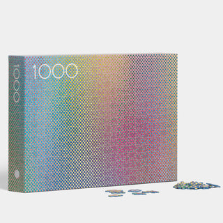 The Play Group’s "1000 Vibrating Colours" 1,000-piece jigsaw puzzle box displays a colourful gradient dot pattern, with several pieces scattered on the white surface nearby.