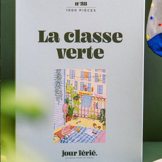 The La Classe Verte | 1,000 Piece Jigsaw Puzzle by Jour Férié features a charming courtyard scene with buildings, plants, and outdoor furniture. Made in France, it offers hours of engaging fun for puzzle lovers.