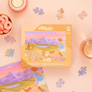 Unwind with the Rockies Meadow 250 Piece Jigsaw Puzzle from Villager Puzzles—a scenic box featuring a woman in a hat, horse, flowers, and mountains—perfect for a relaxing puzzle session with snacks and drinks on a pink surface.