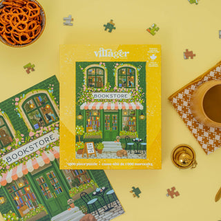 The Emerald Bookstore | 1,000 Piece Jigsaw Puzzle by Villager Puzzles rests on a yellow table with scattered pieces, pretzels, tea, and a gold pocket watch—an ideal gift for book lovers or charming bookish decor.