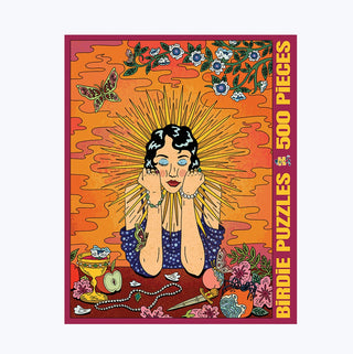 The Holly Holy 500 Piece Jigsaw Puzzle by Birdie Puzzles is made in the USA from premium Blue Kappa chipboard and comes in a colorful box. The design features a stylized woman with sunburst rays, surrounded by flowers, a butterfly, jewelry, and a drink on an orange and red background.