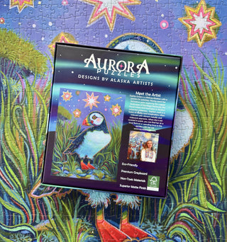 Aurora Puzzles’ Proud Puffin | 1,000 Piece Jigsaw Puzzle showcases an Alaska artist’s puffin illustration, displayed on the finished puzzle against a colorful, starry backdrop.