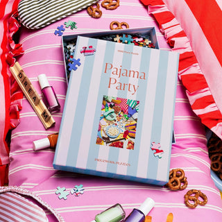 An open Piecework Puzzles Pajama Party | 1,000 Piece Jigsaw Puzzle sits on a pink striped bed, surrounded by nail polish, pretzels, wooden tiles, and colorful recycled paper pieces.