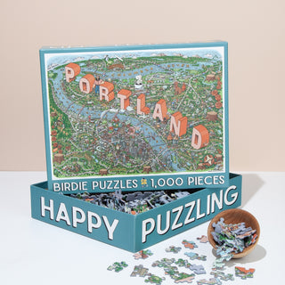 The Portland | 1,000 Piece Jigsaw Puzzle by Birdie Puzzles is shown in its box with loose pieces and a small bowl. The “HAPPY PUZZLING” box makes it an ideal PDX gift for puzzle fans.