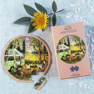 The Van Life | 31 Piece Jigsaw Puzzle by ViSSEVASSE, depicting a whimsical camper van and campfire scene, sits beside its box and a yellow flower on a lace tablecloth—with two pieces missing—creating a charming vignette perfect for your next creative pursuit.